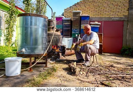 Man Throws Dry Branches Into The Firebox Of A Homemade Distillery Making Moonshine Schnapps, Alcohol