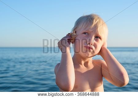 Funny Blond Boy Put Shell To Your Ear Against Sea And Sky Background. Summer Vacation Concept.