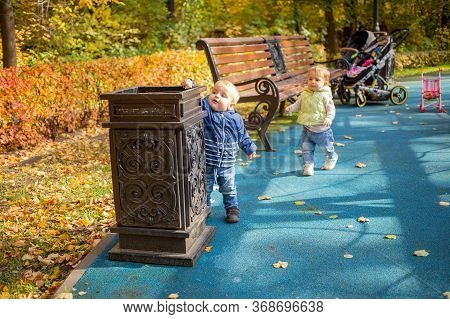 A Little Cute Baby In Blue Clothes Stands Near A Street Urn In A Park And Throws Garbage There And L