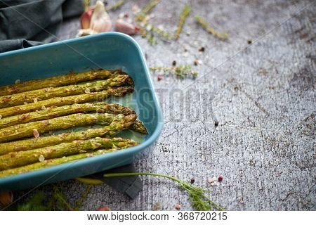 Roasted asparagus seasoned with salt, pepper, garlic and decorated with fresh herbs. Served in blue heatproof dish. Placed on gray stone background.