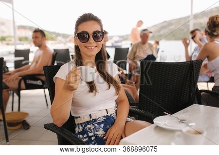 Beautiful Woman Drink Coffe In Cafe Outside. Background Of Sea. Happy Young Woman Sits On Terrace Wi