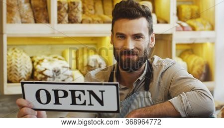 Portrait Of Handsome Smiling Baker Holding An Open Signboard In The Bakery Shop.