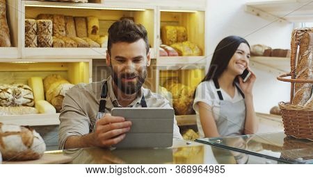 Caucasian Man Vendor Using Tablet Device While Standing At The Counter In The Bakery, His Female Co-