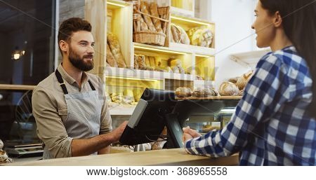 Caucasian Young Man Seller In The Shop Of Bread Talking With Young Beautiful Female Buyer Who Choosi