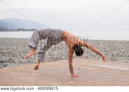 Shirtless Caucasian Man Doing Backflip On Pebble Beach.