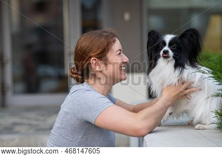Caucasian Red-haired Woman Cuddling With Pappilion Dog Outdoors. Black And White Continental Spaniel