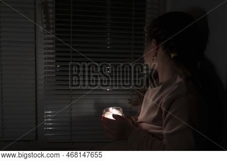 A Girl In A Dark Room Holds A Candle In A Glass Cup And Looks Out The Window. The Concept Of Energy 