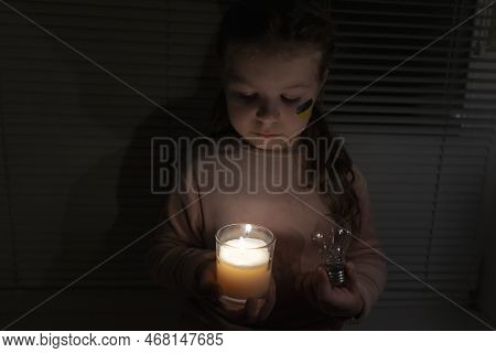 A Girl With A Painted Flag Of Ukraine On Her Cheek In A Dark Room Holds A Candle And An Electric Lig