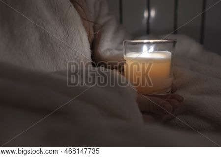 A Girl In A White Plaid Holds A Candle In A Glass Cup In Her Hands Against The Background Of A House