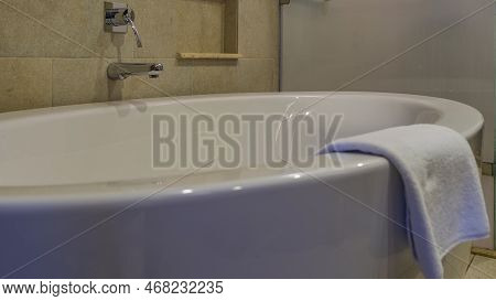Large White Acrylic Bathtub. Close-up. The Towel Folds Around The Edge. A Shiny Metal Faucet And A S