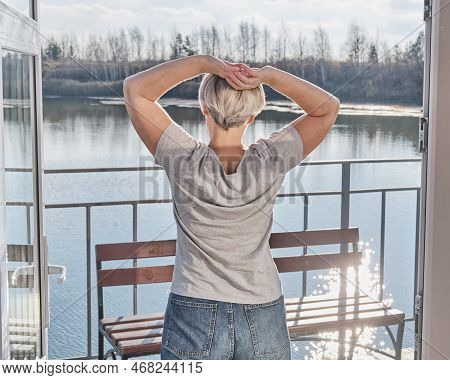 Senior Woman With Short Gray Hair Stretching Oneself On Veranda Of Country House On Lake Shore. Enjo