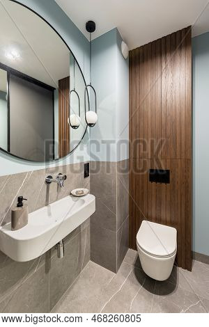 Modern Bright Bathroom With White Toilet Seat On Wooden Lamela Wall. Round Mirror With Black Frame.