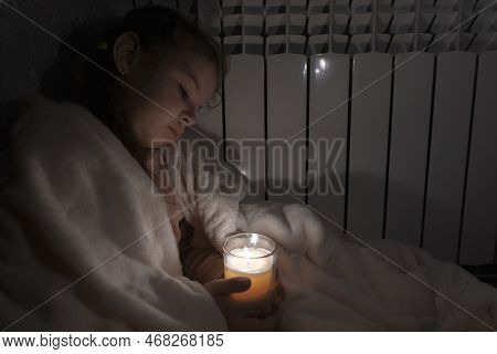 A Girl In A White Plaid Holds A Candle In A Glass Cup In Her Hands Against The Background Of A House