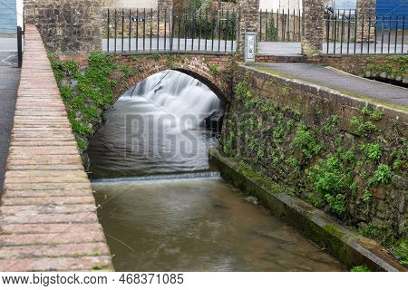 Long Exposure Of A Watefall Flowing Under A Bridge On The River Lim Walkway At Lyme Regis In Dorset.