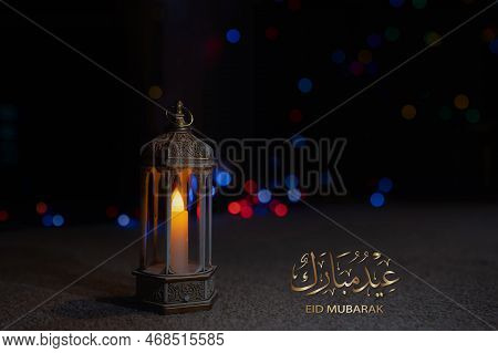 Eid Mubarak Calligraphy Greeting Design With Islamic Lantern On Carpet With Blurry Light Background.