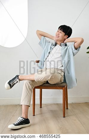 Happy Asian Man In Casual Clothes Sits On A Chair Relaxing In His Minimal Bright Living Room.