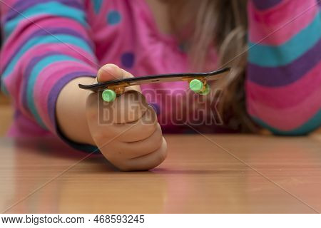 A Girl Holds A Fingerboard In Her Hand While Sitting At A Wooden Table, Close-up, Selective Focus.