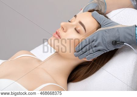 A Cosmetologist Doctor Makes A Microcurrent Massage Procedure On The Face And Neck Of A Young Beauti