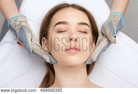 A Cosmetologist Doctor Makes A Microcurrent Massage Procedure On The Face And Neck Of A Young Beauti