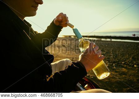 A Man On The Shore Of The Sea Or Ocean Opens A Bottle Of White Wine. A Date On The Beach, Alcohol On