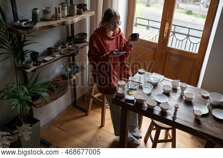 Young Woman Craft Artist Sitting At Table At Cozy Home Ceramic Studio, Using Smartphone And Drinking