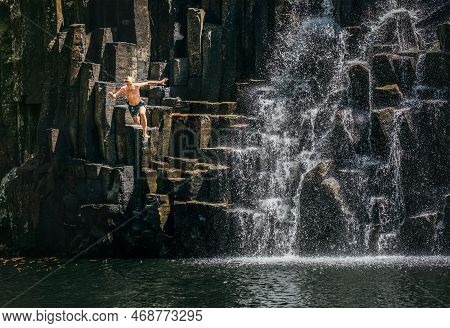 Teenage Boy Jumping In Waterfall Lake. Falling Water Streams Flow On Black Volcanic Stone Cascades. 