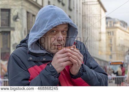 A Homeless Man 45-50 Years Old In A Jacket And Hood With A Mug In His Hands On The Street, Close-up.