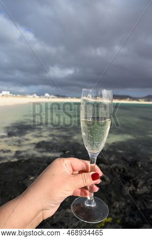 Hand With Glass Of White Champagne Or Cava Sparkling Wine On White Sandy Tropical Beach And Blue Oce