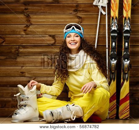 Happy woman with skis and ski boots sitting on a floor near wooden wall