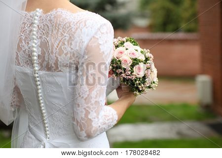 Bride in a beautiful white wedding dress with lace and pearls stands with her back and holds a wedding bouquet of roses in her hands. Horizontal shot daylight summer.