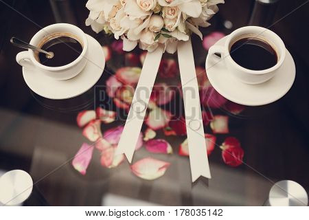 A beautiful composition with engagement rings two cups of coffee a bouquet of flowers ribbons and rose petals on a glass table