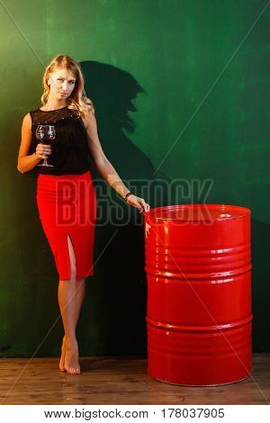 Woman in Red Skirt Stands Near the Barrels of oil. Sexy Attractive Blonde Holds Glass of Wine and Looks to Camera.