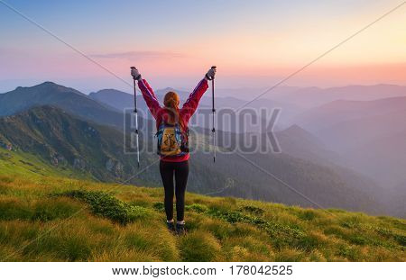 Redheaded girl athlete with a backpack and sticks stands on the green hillocks and looks at high mountain landscapes and fascinating sunset.
