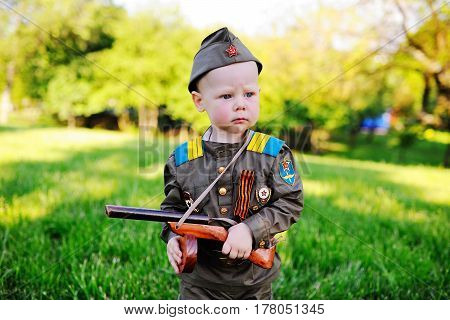 A cute child in the uniform of a Soviet soldier with a St. George ribbon with a toy gun on the background of nature. May 9, February 23, defender, against fascism.