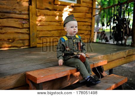 Child in military uniform with a toy gun on the background of a wooden house
