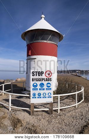 Old red and white lighthouse and building site warning sign close to Assens new harbor bridge in Denmark.