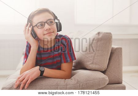 Teenager enjoy music - portrait of dreamy kid in headphones while lying on sofa indoors. Relax time - teenage boy listen to music on couch at home