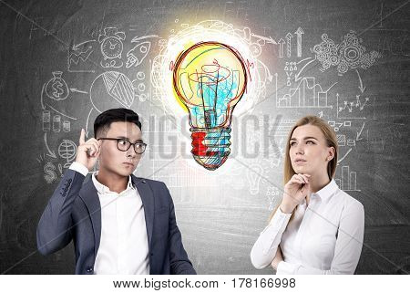 Portratif of a blond businesswoman and an Asian businessman standing near a blackboard witha a large and colorful light bulb drawing on it.