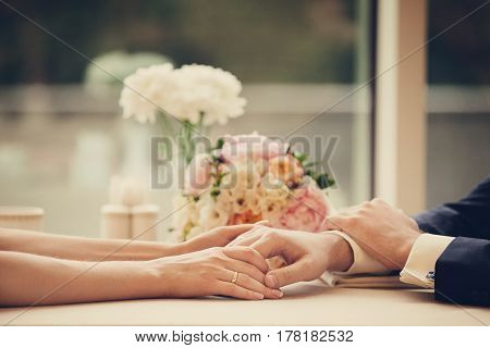Married couple holding hand each other while sitting by table in cafe, on background of bouquet of beautiful flowers. Closeup