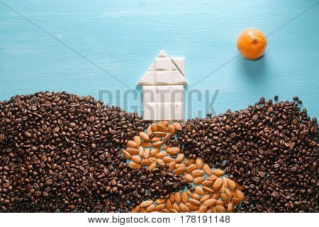 The house from white chocolate, the earth from coffee grains, the road from almonds, the sun from a citrus on blue background. Abstract symbols flat lay composition. Concept of realty, cosiness, comfort, happy family etc.