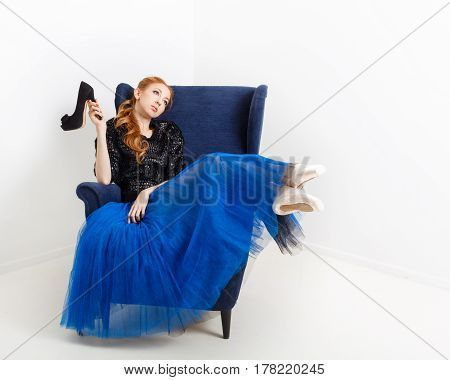 Pretty ballerina in tutu and pointe shoes sits in a chair. She holds high-heeled shoes.