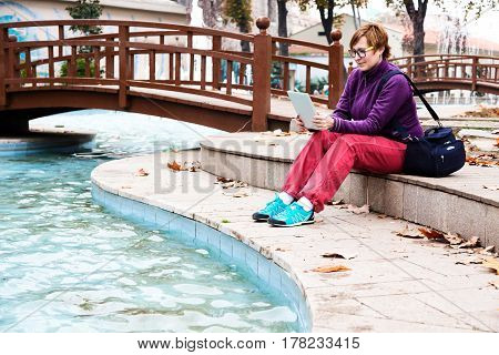 Cute Woman in casual and sporty Clothing sitting in Park next to Pond and using tablet pc device