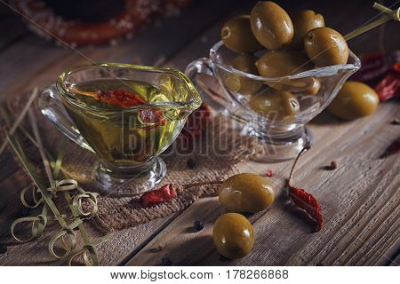 Premium extra virgin olive oil and green olives with fresh herbs black pepper and chili pepper on rustic wooden background.