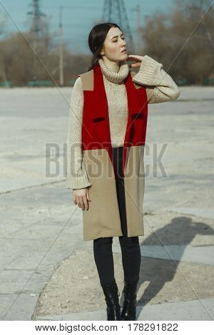 Young beautiful girl model in vest coat of red and beige color and in turtleneck skinned. She also wears dark trousers. The hair of a woman is braided into the tail, and curls weigh in front.