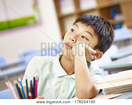 eleven year-old asian primary schoolboy sitting in classroom alone dazing or daydreaming.