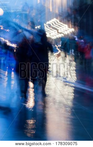Abstract background in naturale blue tones. People walking down the city street in rainy evening. Light illumination from lanterns and shop windows. Intentional motion blur. Concept of seasons, weather, modern city.