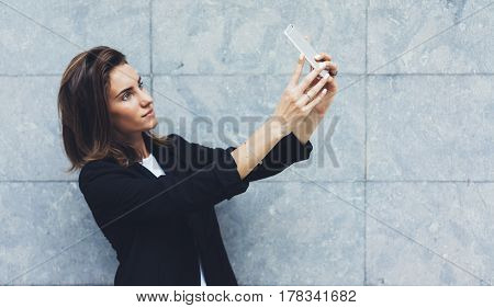 Portrait young businesswomen in black suit using smartphone isolated on background concrete gray wall mockup pretty hipster manager holding mobile gadget girl making selfie photo connect