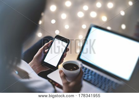 Hipster girl using computer and cup coffee in home atmosphere person holding blank smartphone on background glow illumination female hands texting on relax glitter bokeh mockup templates blur
