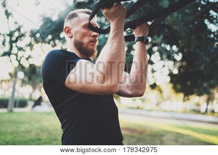 Muscular athlete exercising trx push up outside in sunny park at the morning.Attractive fittness man doing workout exercises outdoors.Blurred background.