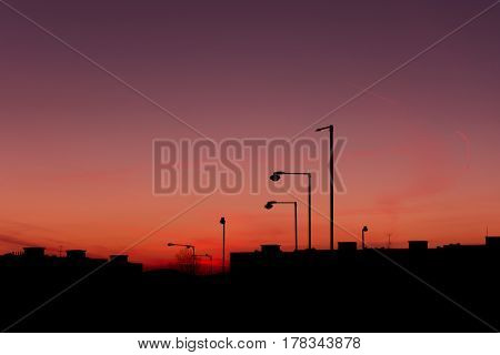 Silhouette of buildings and streetlights in city before sundown.  The sky is red and purple with alpenglow. Evening in city